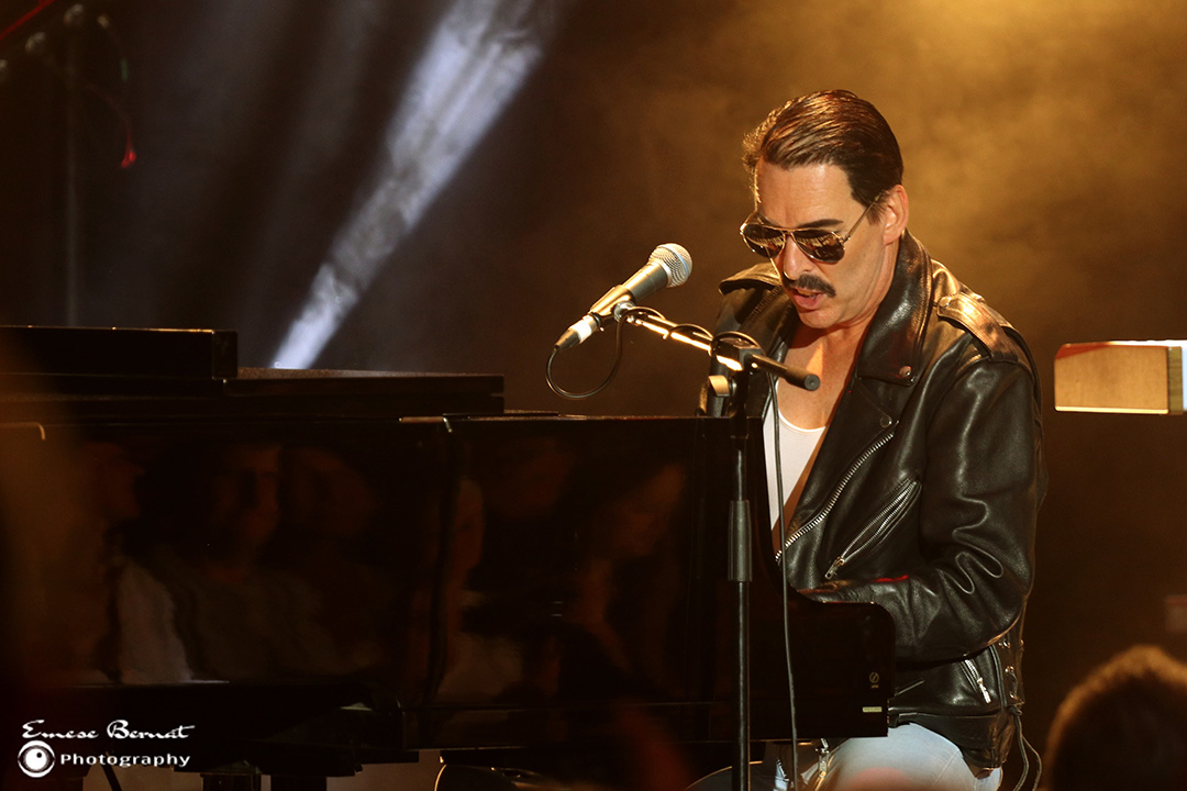 A man waering a black leather jacket and aviator sunglasses sits at a grand piano