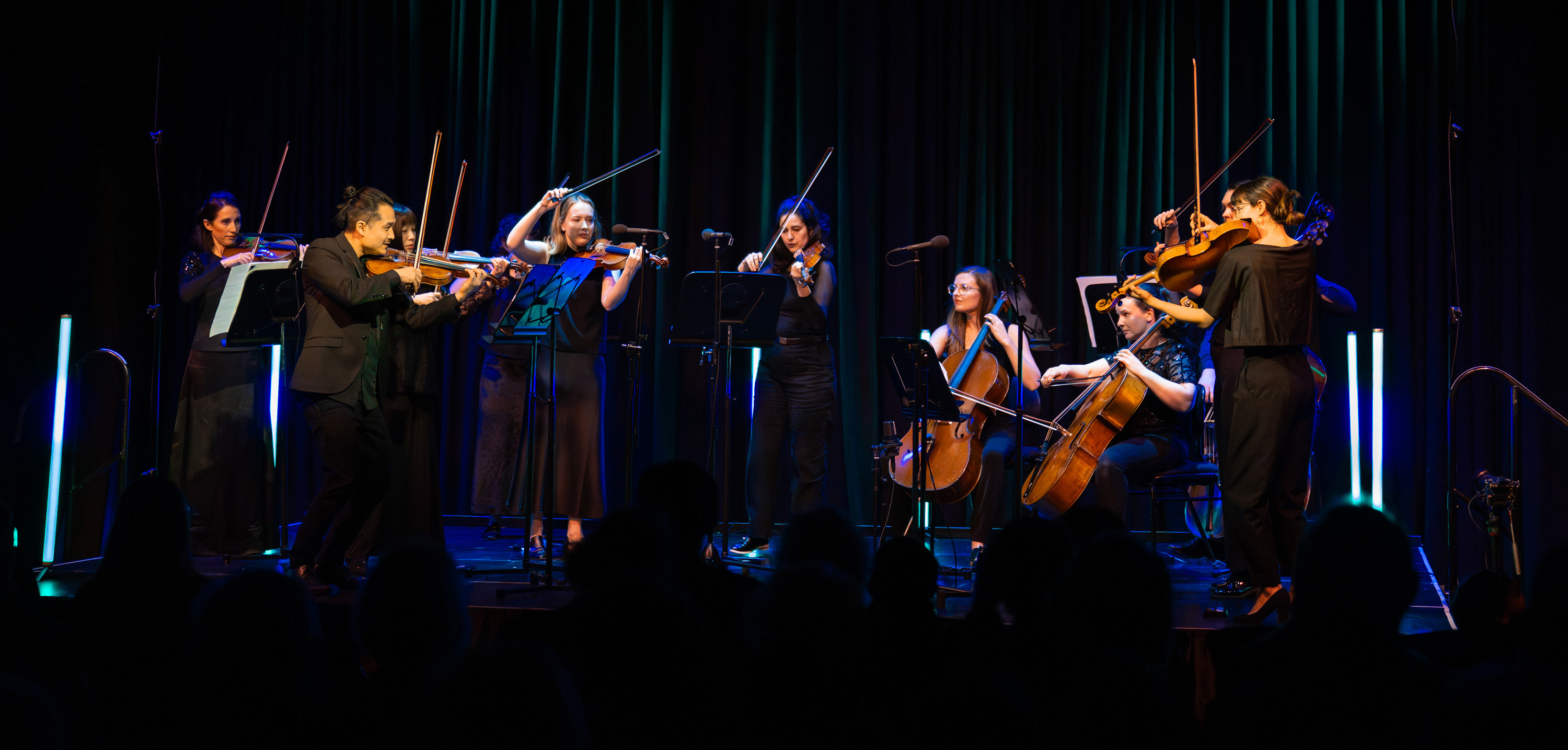 A group of musicians performs onstage under blue and purple lighting, with violinists, cellists, and other string players playing together. Audience silhouettes fill the foreground.