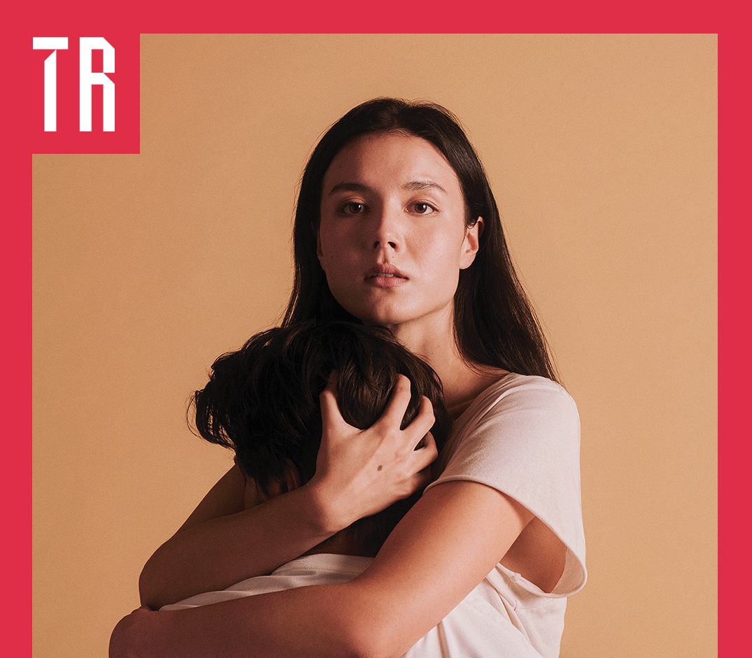 Photo: Pierre Toussaint A couple stand against a neurtal tan coloured background. A woman embraces a second person who's face is truned away from us.