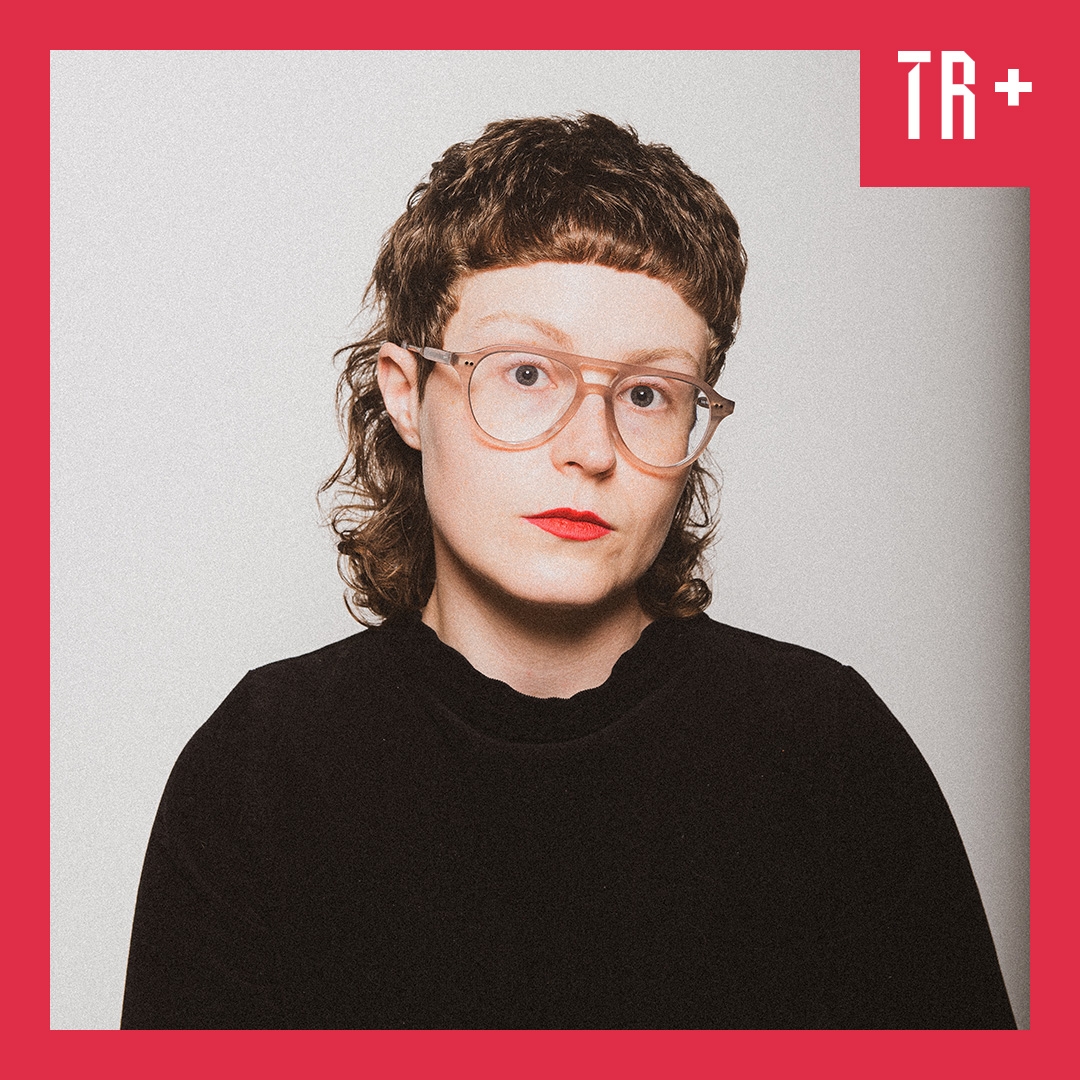 Photo: Nick Robertson A portrait of non-binary comedian Scout Boxall. They are wearing a black jumper, glasses, bright red lipstick.