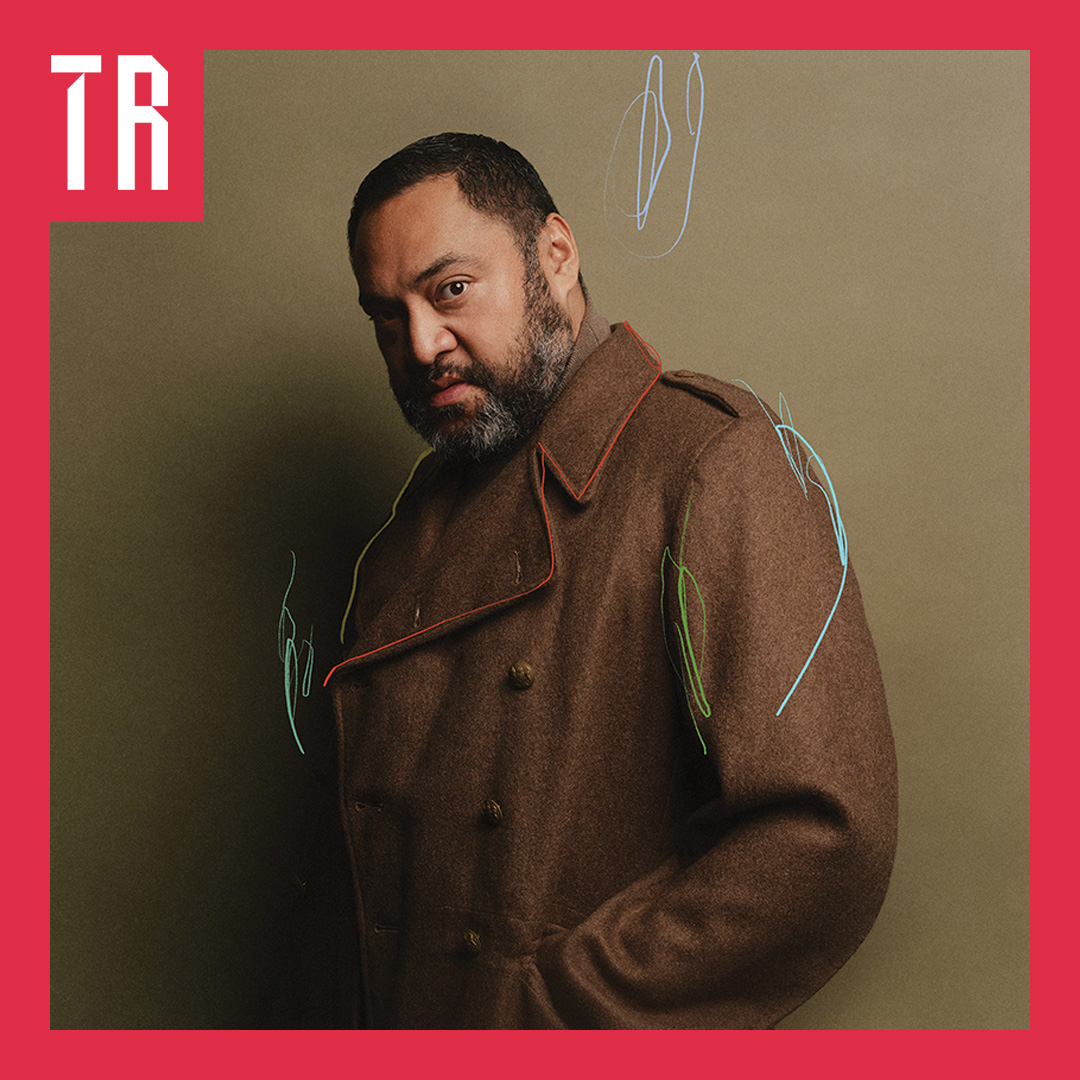 A heavy set man wearing a brown trench coat and beard looks directly to camera