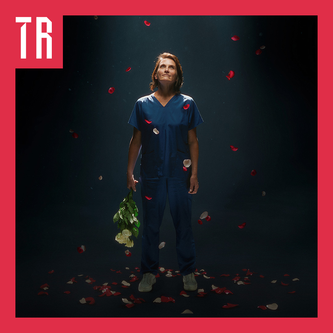 A woman stands against a dark background, she is wering blue hospital scrubs and is holding a bunch of flowers. Petals fall from above her.