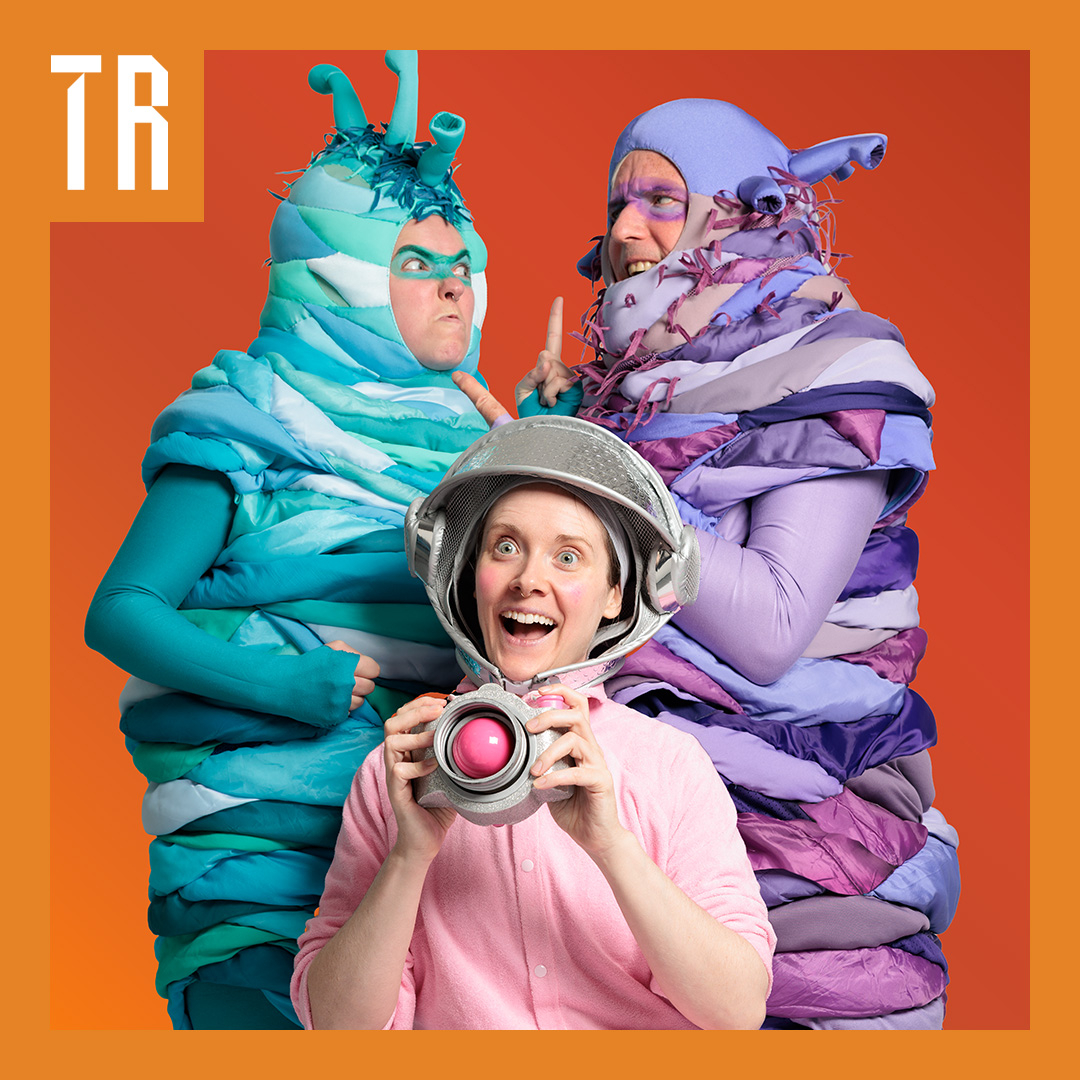 A colourful photo of three performers dressed in costumes, two a dressed as worm like aliens and one like and explorer.