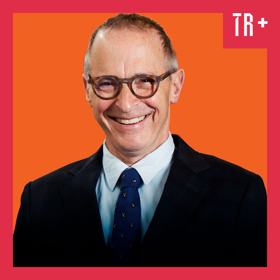 A portrait of writer and humorist David Sedaris standing in a library, wearing a blazer and tie, holding a pipe