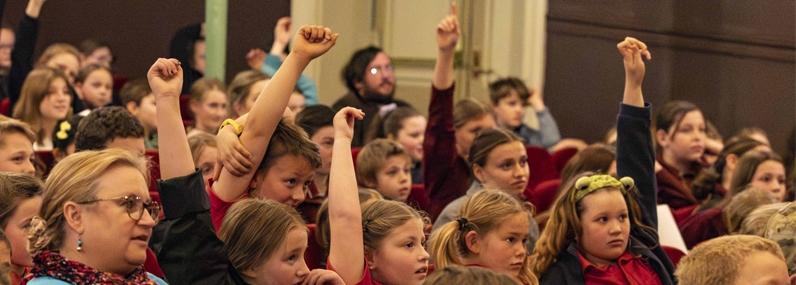 Photo: Jillian Mundy A group of students sit in the red theatre seats. Some of them have their hands raised waiting to ask a question.