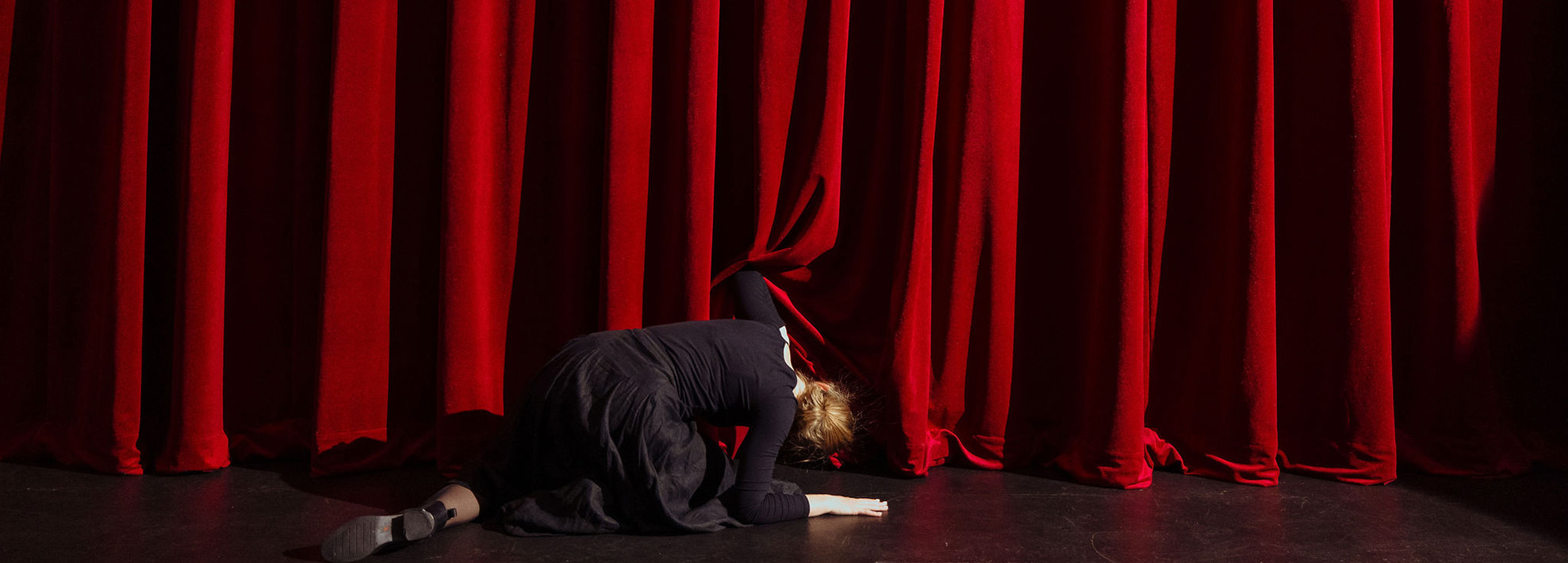 A woman wearing a black dress peaks under a red velvet theatre curtain