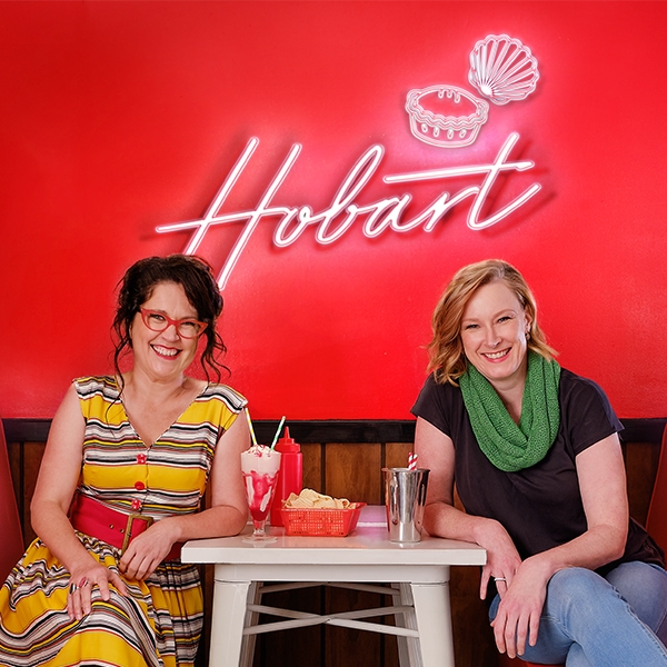 Chat 10 Looks 3 Live Two women sitting in front of a red wall with a neon light that says Hobart. They are sitting at a table with milkshakes. The woman on the left is Annabel Crabb and the woman on the right is Leigh Sales.