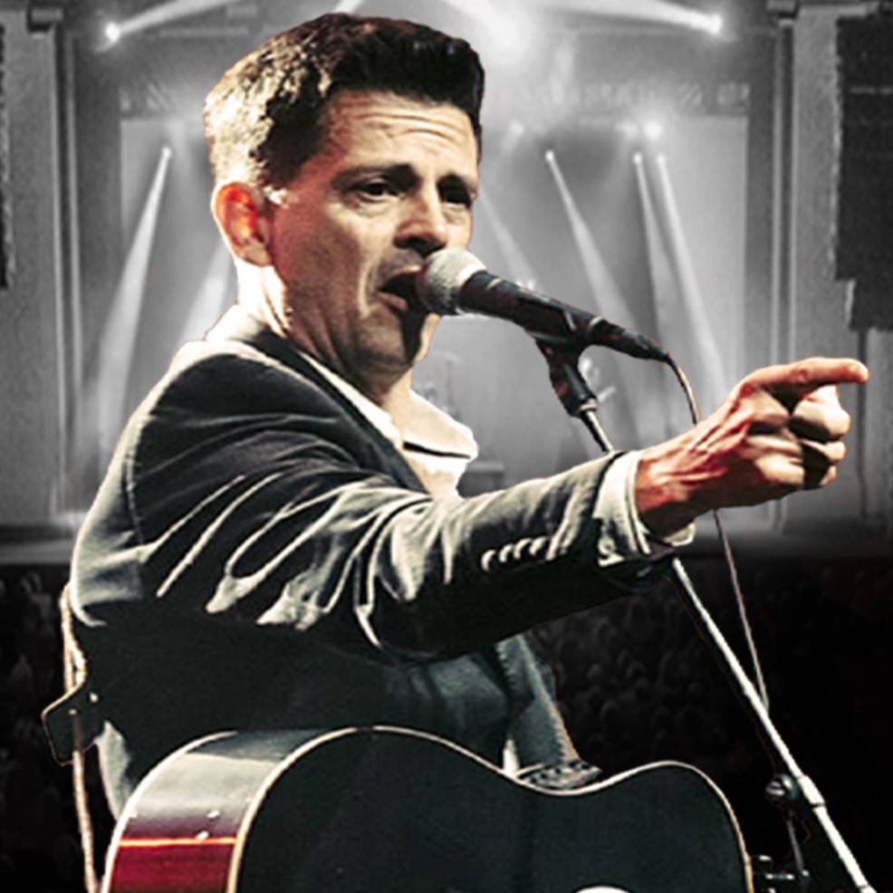 A man impersonating Johnny Cash. He is wearing a black suit jacket and a white shirt. He is holding a black guitar and he is singing into a microphone. He is pointing into the audience.