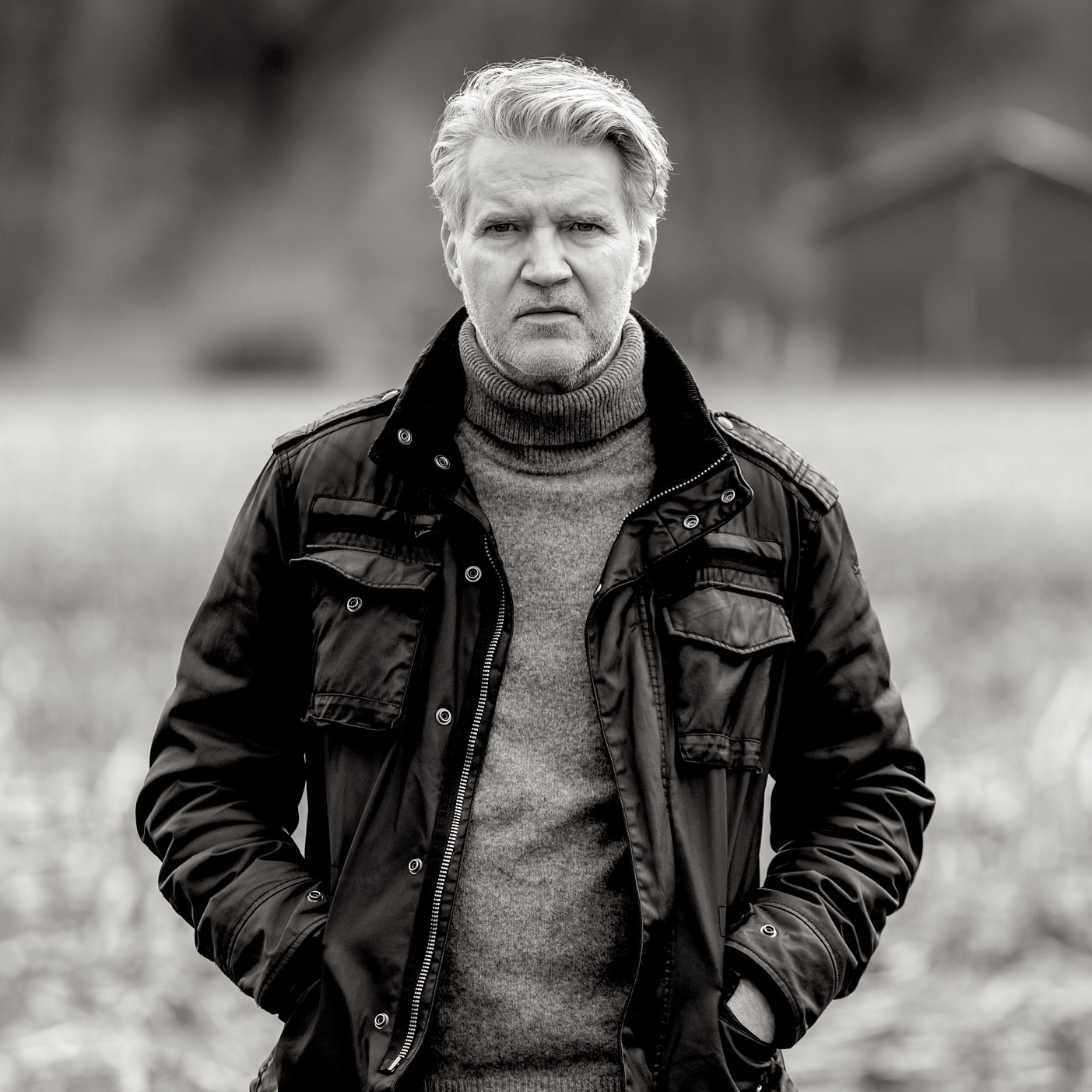 Lloyd Cole A black and white photo of a Lloyd Cole in a field. He is wearing a black jacket and a grey jumper underneath. He has his hands in his pockets and he is looking into the camera.