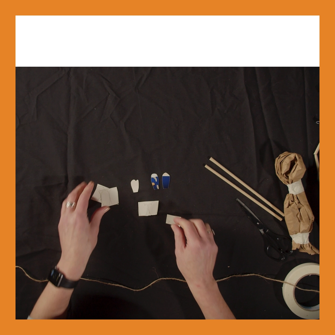 A pair of hands seen from above at a table. The table has a black cloth over it and the hands are making something from paper. There is an orange border on the image.