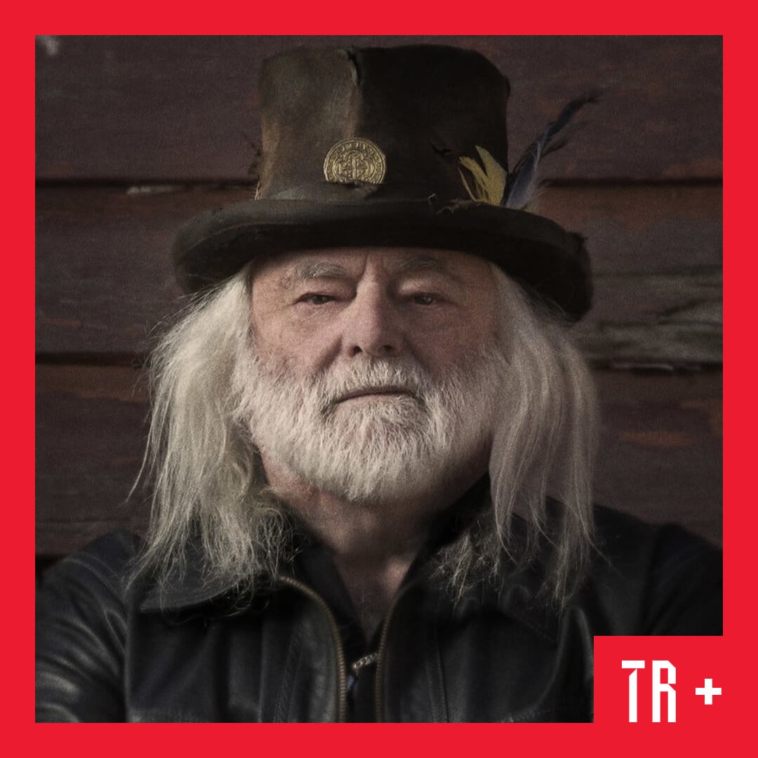 Brian Cadd wearing a leather jacket and top hat with feathers pined to its side. He has white long hair and a beard.