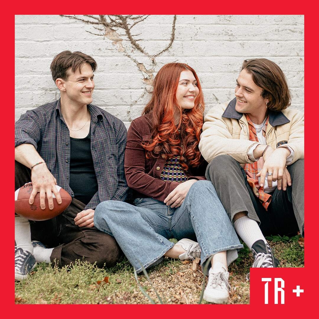 Image: Joshua Howells - GOLDINGcreative Three young people sit on the ground in front of a whitewashed wall. They are looking adoringly at each other.