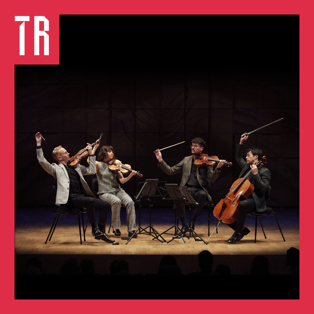 Four musicians are seated on a stage. They are all playing stringed instruments. They are waving their bows in the air. There is a spotlight on them. .