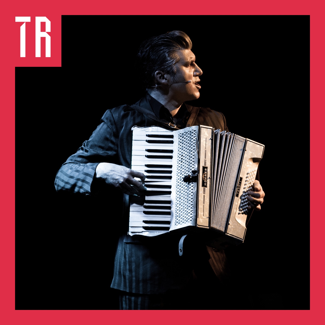 A man holds a piano accordion and looks to be playing it. His face is turned to the right.
