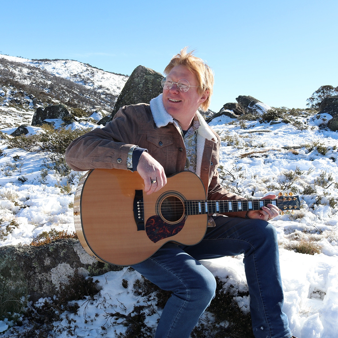 Darran Coggan A man with light red hair and glasses wits with a guitar in an alpine wilderness