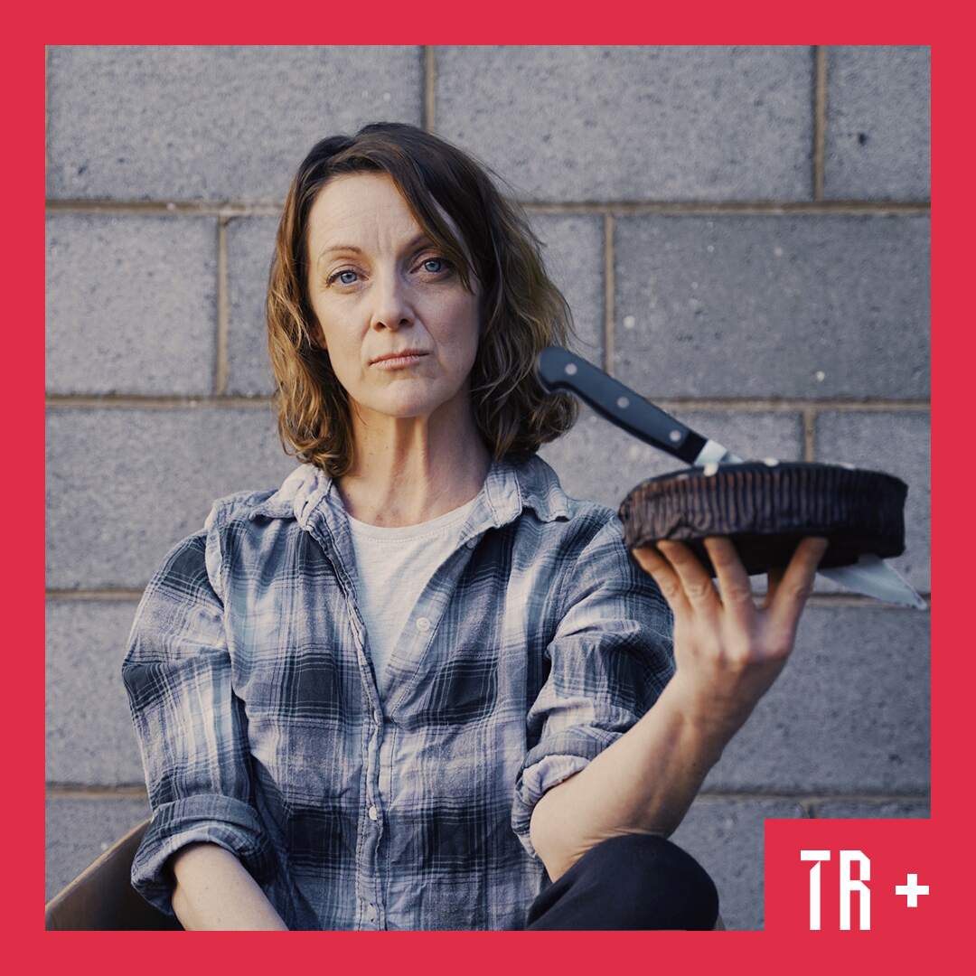 Photo: Luca Schild A woman holds a cake with a knife stabbed into it. She looks directly at the viewer with an intense stare. She is standing in front of a concrete brick wall.