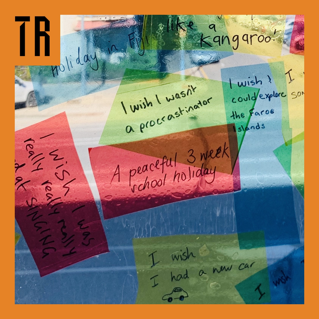 Colourful pieces of cellophane are stuck to a window. They have wishes written on them in black texta. They overlap.