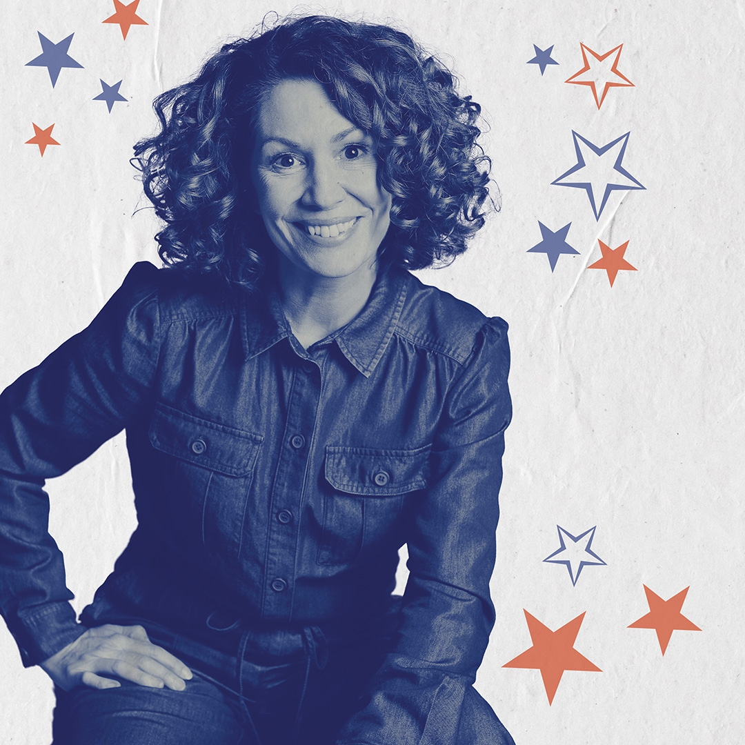 Kitty Flanagan live A lady with big curly hair is sitting wearing a jumpsuit. She rests her hand on her leg, and is smiling
