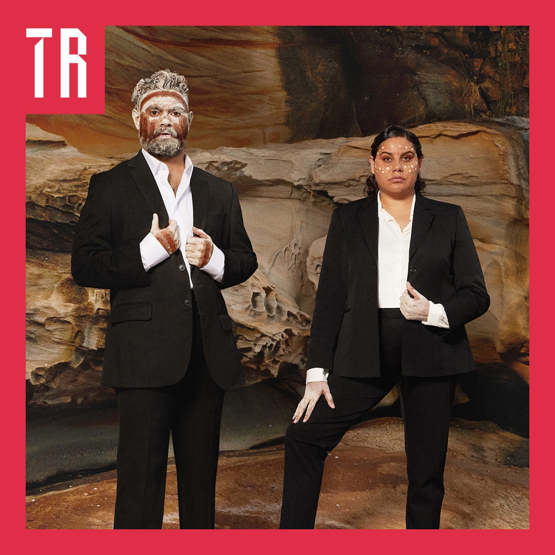 The Visitors Two First nations people, one male one female, stand infront of a sandstone cliff. They are wearing black business suits and are adorned in traditional markings. Their pose is confident and powerful.
