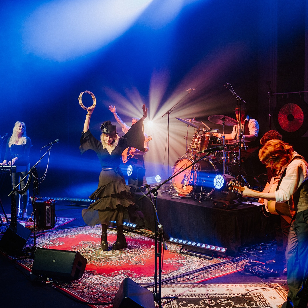The Fleetwood Pac A live band perform on a stage, they have bohemian rugs on the floor and are dressed in boho clothes