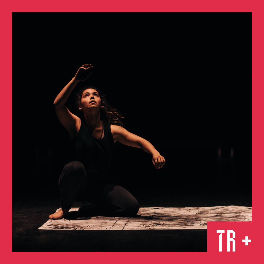 Photo: Grace Chia A First Nations performer looks to the sky her right arm raised up. Light shines on her from above. She is crouched on a charcoal marked piece of paper.