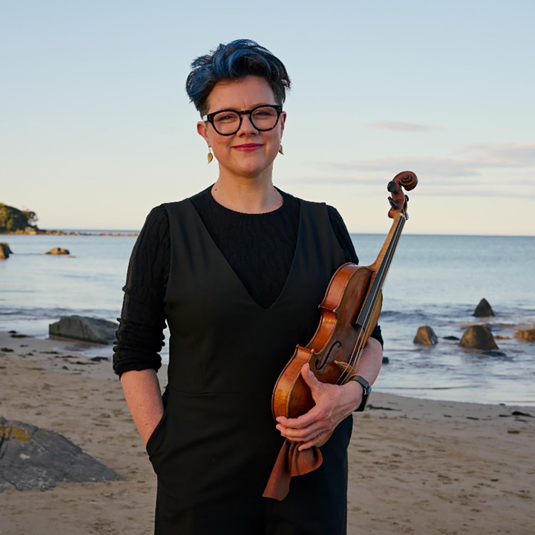 Julia Fredersdorff HERO image A woman with a pixie hair cut and dark rimmed glasses stands on a Tasmanian beach, she is holding a violin in her hands and smailing gently