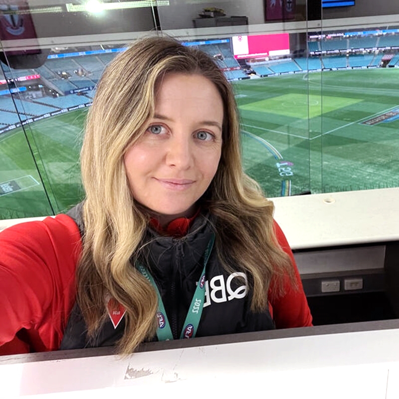 Michelle Cooling A woman with long wavy blonde hair has taken a selfie with a soccer pitch in the back ground. She is in a box seat