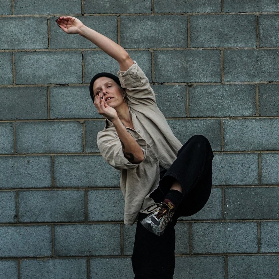 Danceweekender hero A female dancer in baggy clothes moves gently against a cold grey brick wall