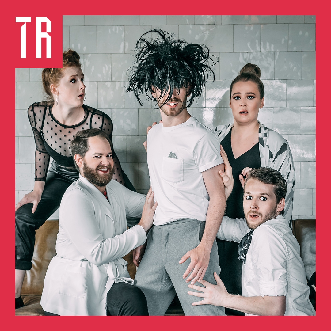 A group of 5 performers stand in mock suprose. The central figure is wearing an upside down brown wig