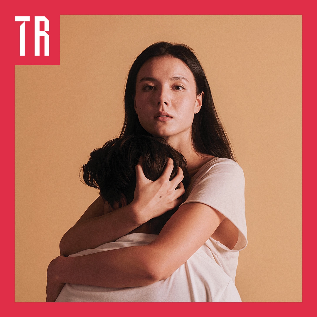 Photo: Pierre Toussaint A couple stand against a neurtal tan coloured background. A woman embraces a second person who's face is truned away from us.
