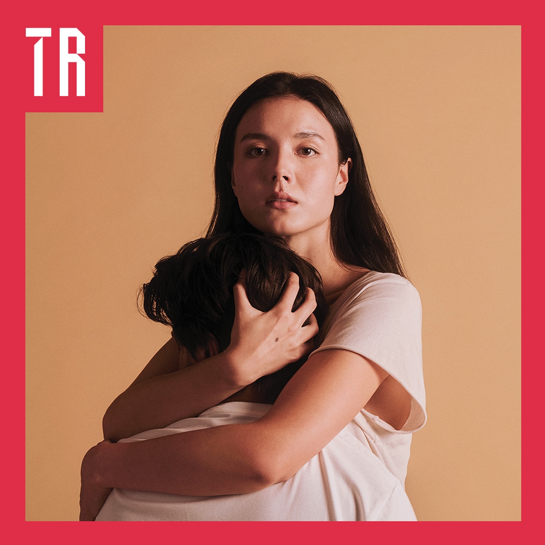 Photo: Pierre Toussaint A couple stand against a neurtal tan coloured background. A woman embraces a second person who's face is truned away from us.