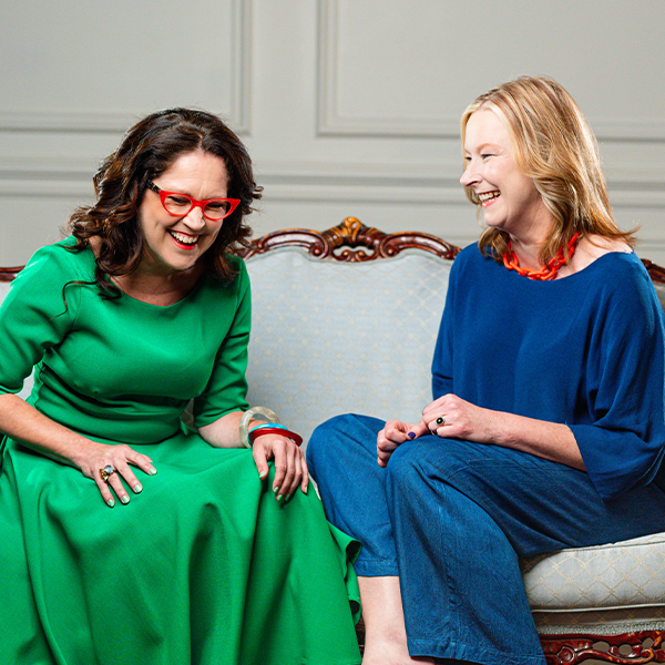 Two woman, each wearing bold colours, sit smiling and laughing on a couch