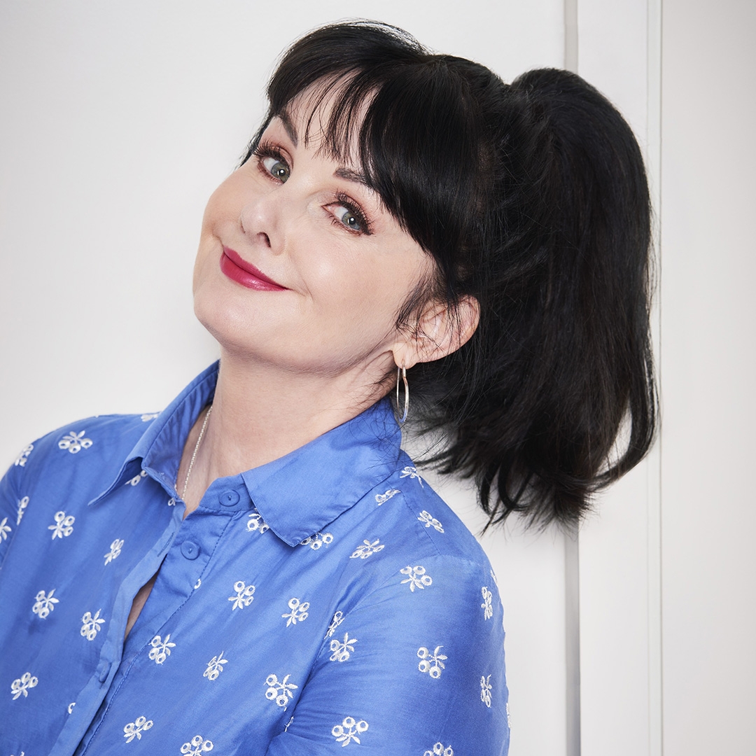 A woman with dark hair and pale skin is smiling in a bright blue shirt