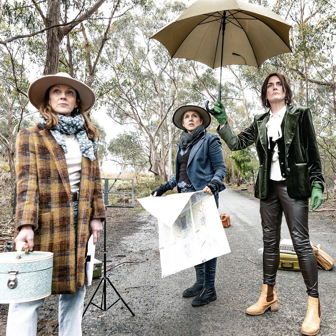 Three woman stand on a road. One holds a case, one a map and one an umbrella. They are all looking out to the distance.