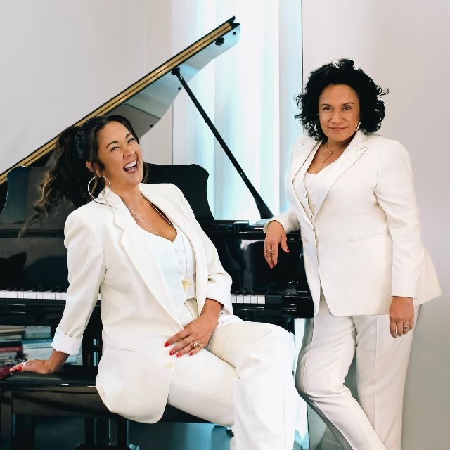 Photo Lisa Businovski An Evening with Vika and Linda hero image. Two sassy women dressed in white casual suits lounge around a black grand piano