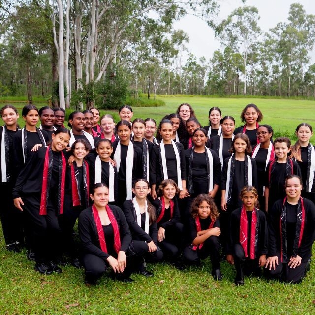 Gondwana Childrens Choir hero A large group of children dressed in black with red and white scarfs stand and crouch on a green park land surrounded by gum trees