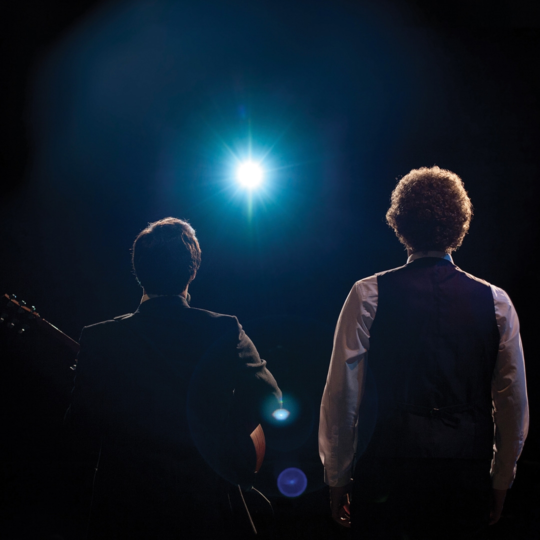 Simon and Garfunkle hero image Two male figures stand looking out into a stage light