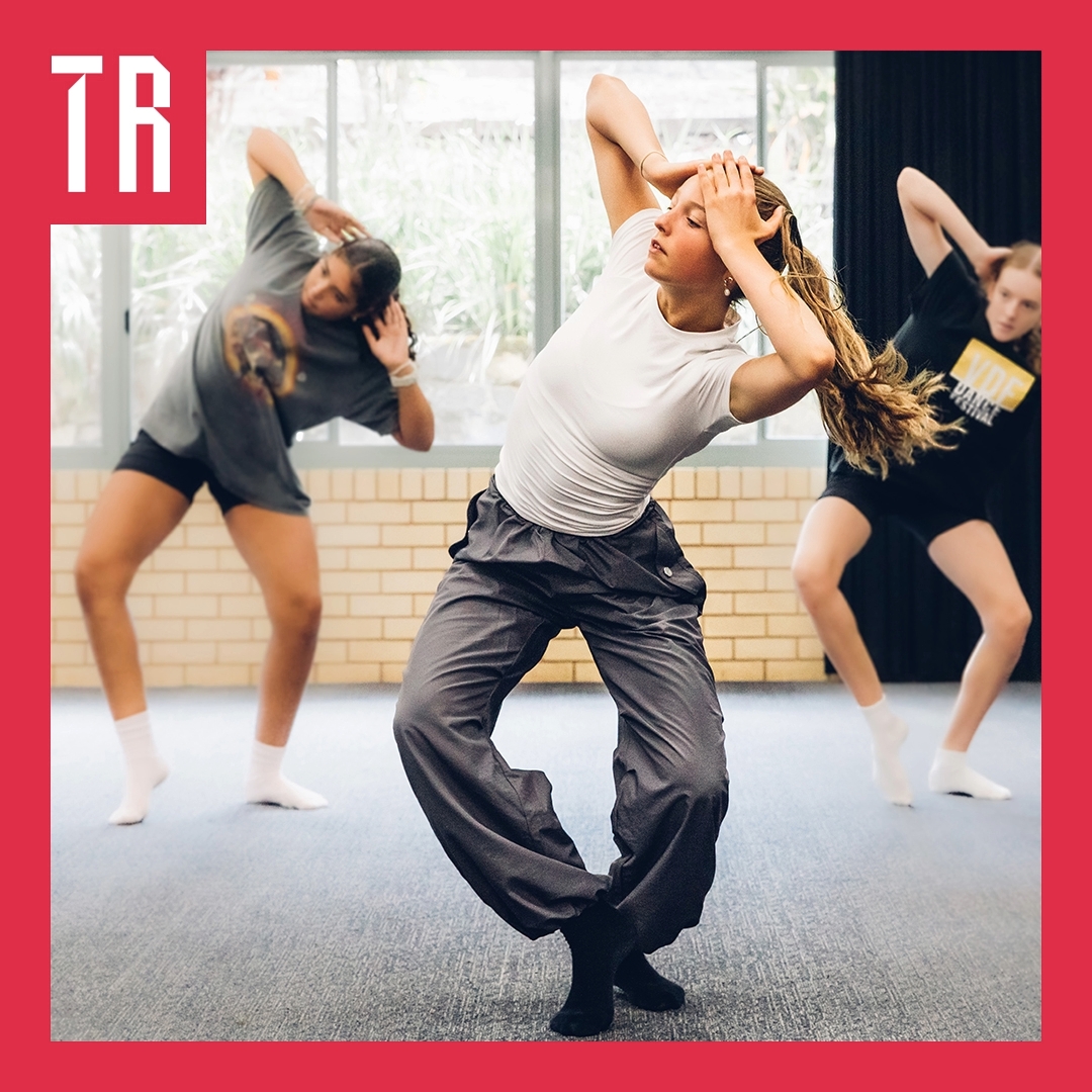 Photo: K Holmes Three dancers are in a carpeted room with windows behind them. They are doing a synchronised move with both hands on their heads.