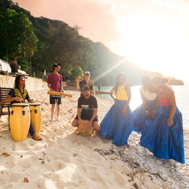 Hero image Salama Maloya Culture Night - Island Rhapsody A group of people stand on a tropical beach. There are male and female musicians and dancers