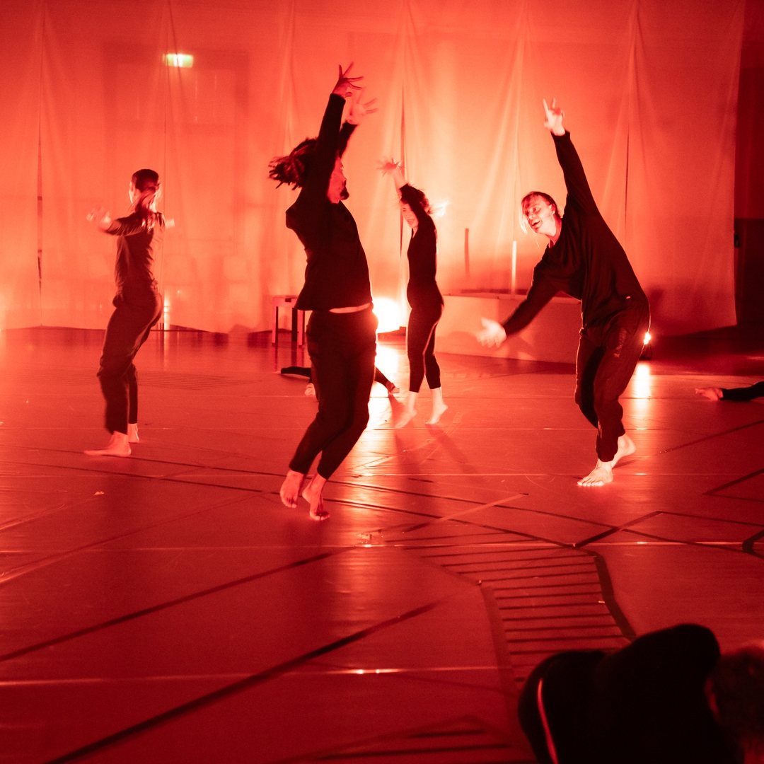 Photo: Amy Brown A group of dancers jump in the air with their arms outstretched. There is red light and they are wearing black clothing.