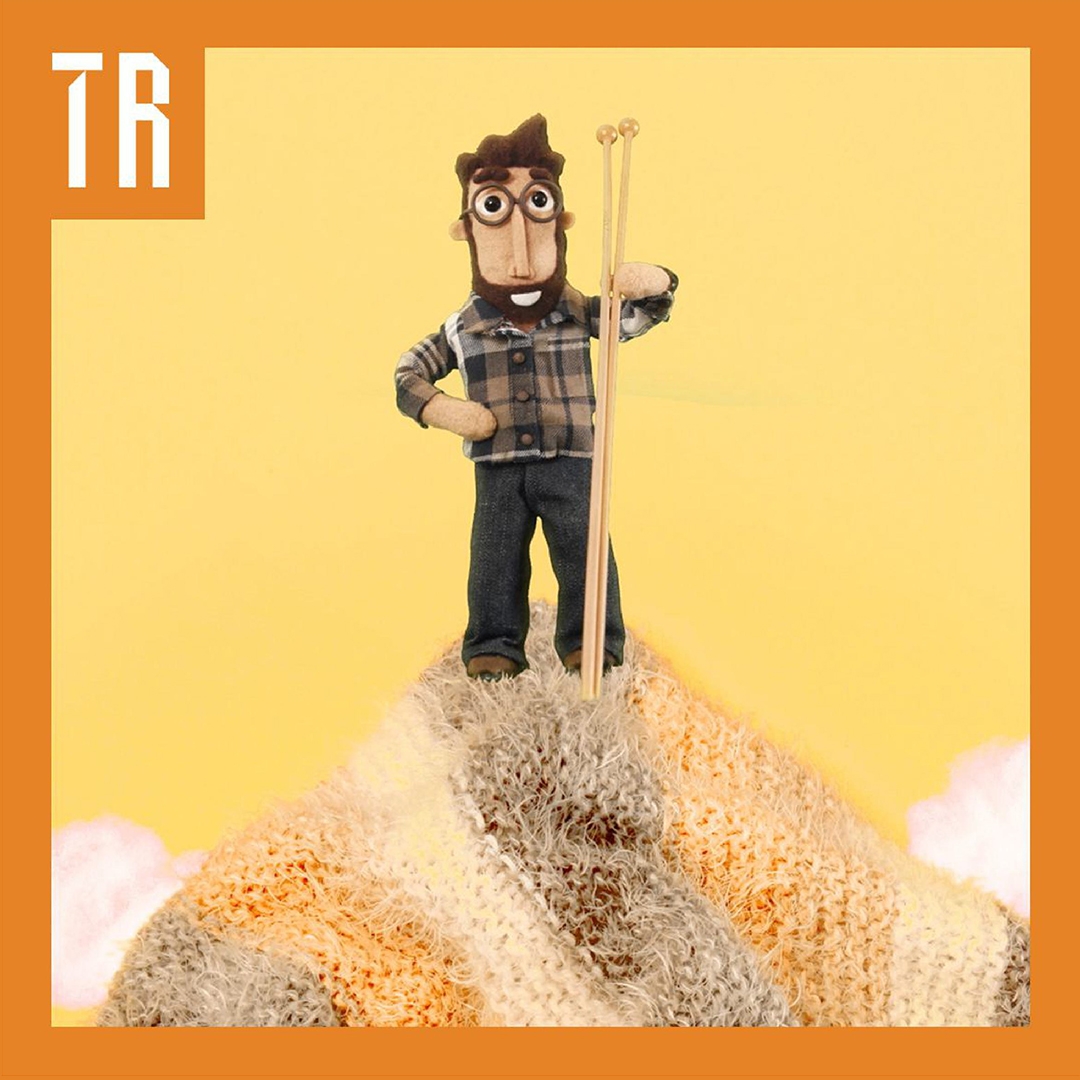 A felt man stands upon a mountain made of knitted fabric. He holds knitting needles which are the same size as him. He has a beard and wears glasses and is dressed in a checked shirt and grey pants.
