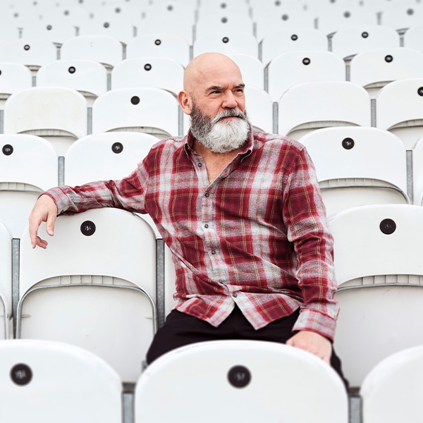 Image: supplied A man with a bald head and grey beard, wearing a red flannelett shirt sits in white auditorium stalls