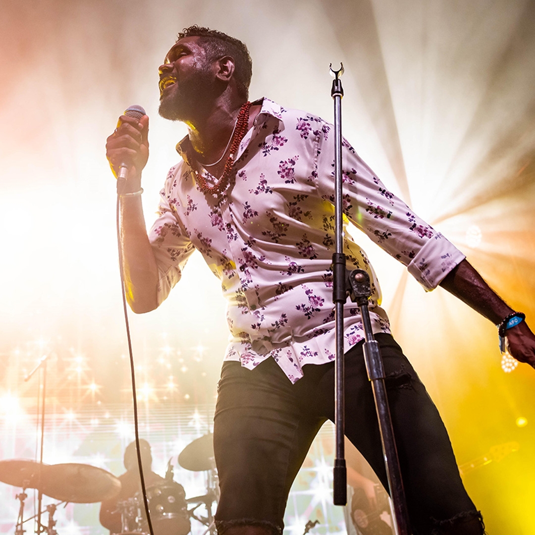 Image: Lachlan Douglas A man holds a microphone on stage, he is joyously singing. Golden light radiates like a sun behind him