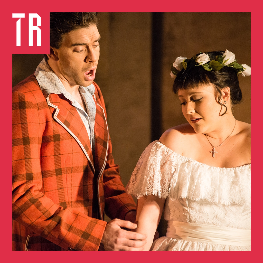 A man waering a red tweed coast sings to a woman in a white lace off the sholder dress with white roses in her hair