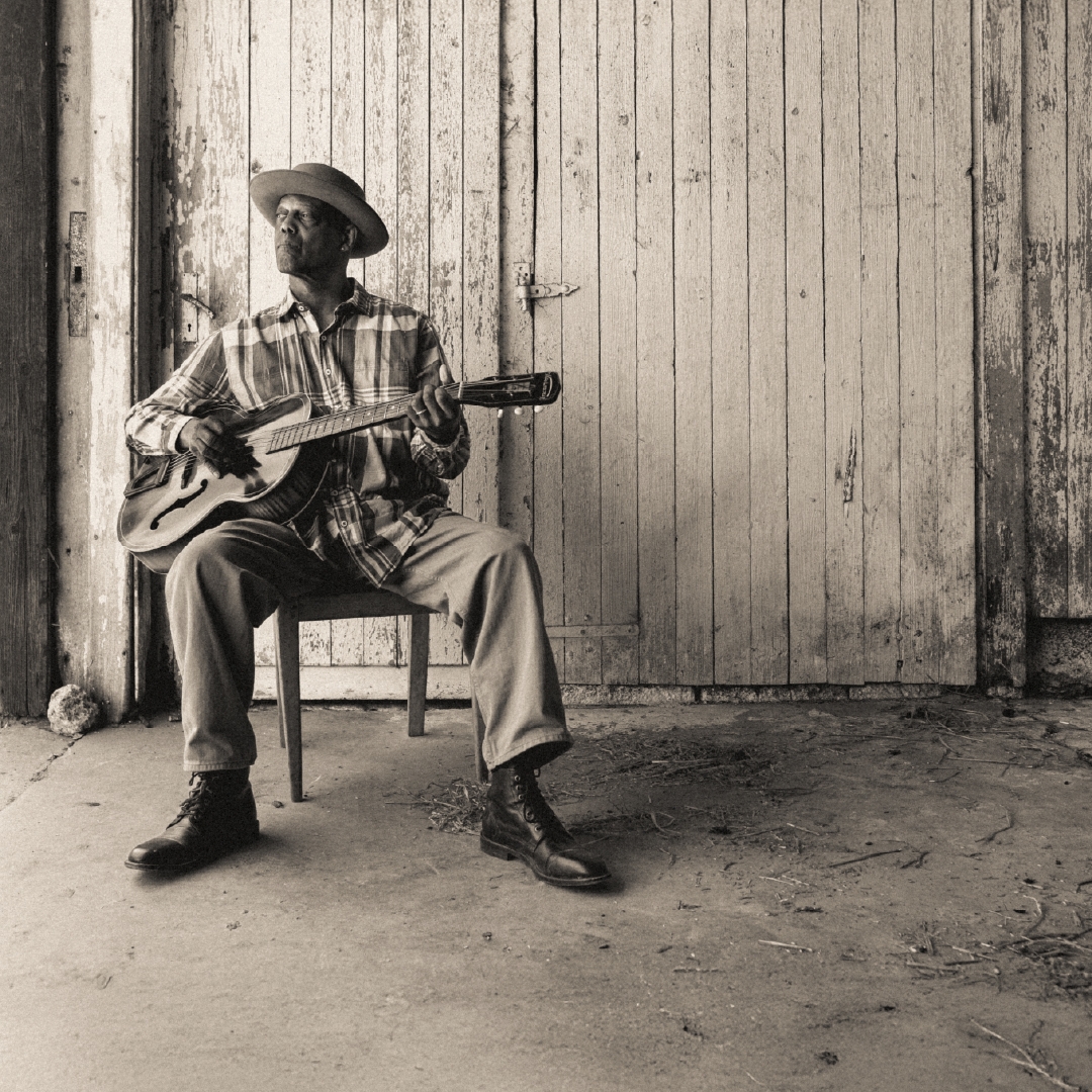 An African American man leans back on a wooden chair, a guitar rest gently under his arm as he strumms it.