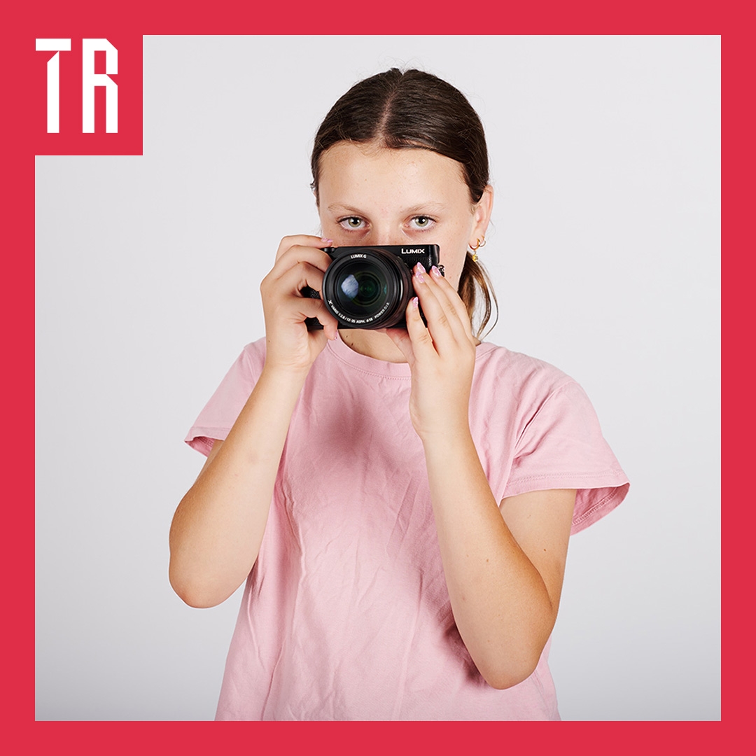 A teenage girl holds a camera in front of her face, her eyes can been seen over the top of the camera looking directly at us