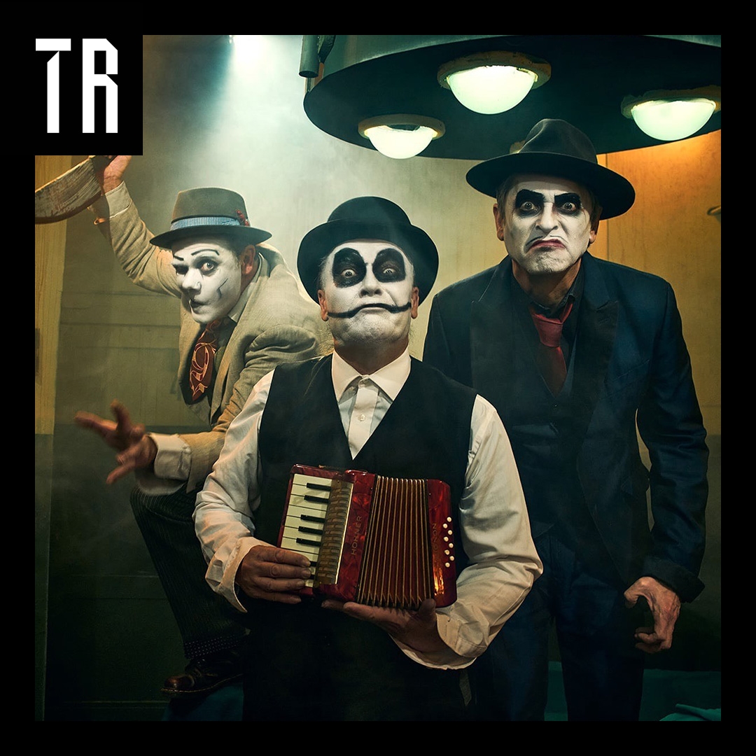 Three musicians pose wearing skull face paint , 1930s gangster attire and fedoras. The man at the front holds a piano acordian.