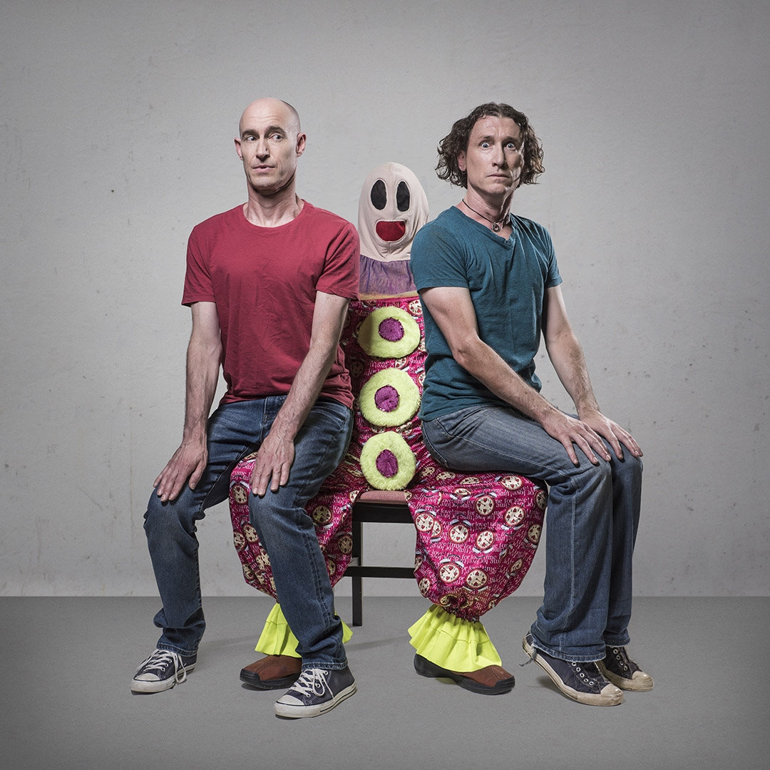 Two men are sitting back-to-back on a whimsical, human-sized puppet figure that is acting as a bench. The puppet has a large, cartoonish face with black eyes and a red mouth, and it is covered in bright, colorful fabric with circular patterns. The man on the left is bald, wearing a red shirt and jeans, while the man on the right has curly hair, wearing a teal shirt and jeans. Both men have serious expressions and are sitting upright with their hands on their knees. The background is plain and neutral, drawi