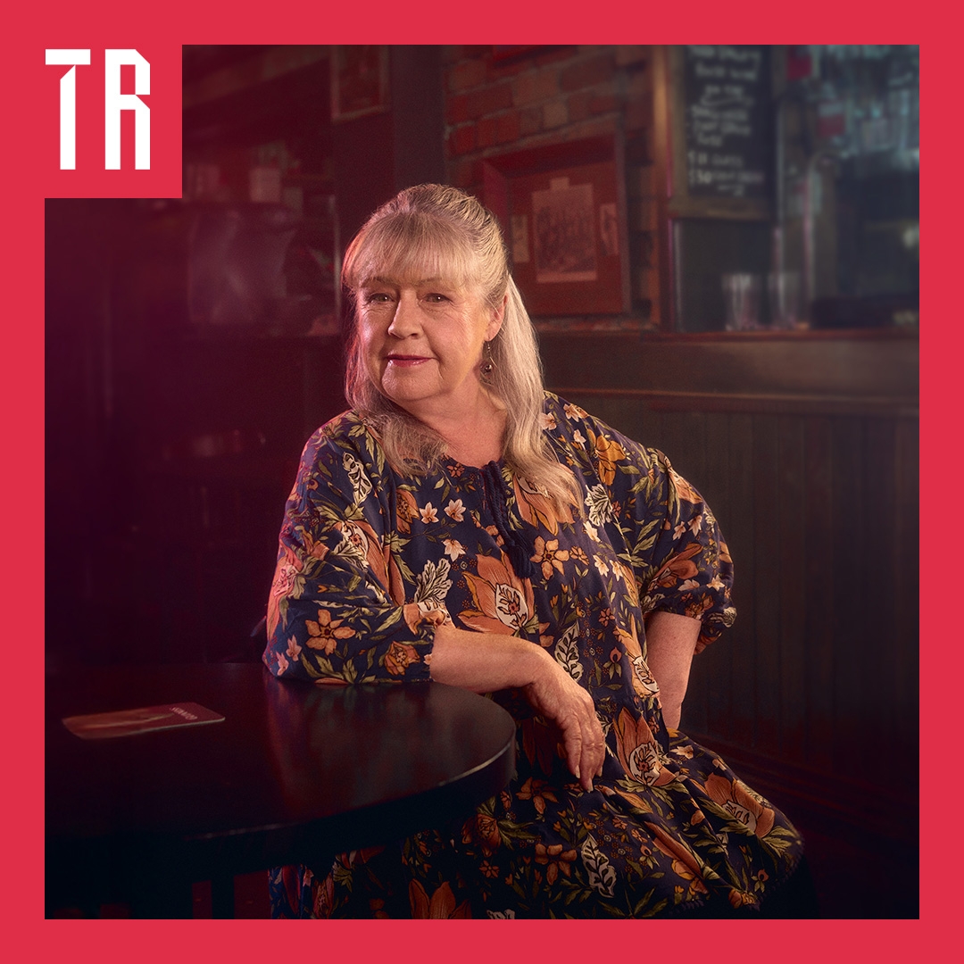 A woman sits on a chair, leaning her arm on a dark woodern table. She is smiling, in a wise way. She wears a floral dress of black and tan colours. Her hair is greying blonde. She look as if she is about to tell us a story