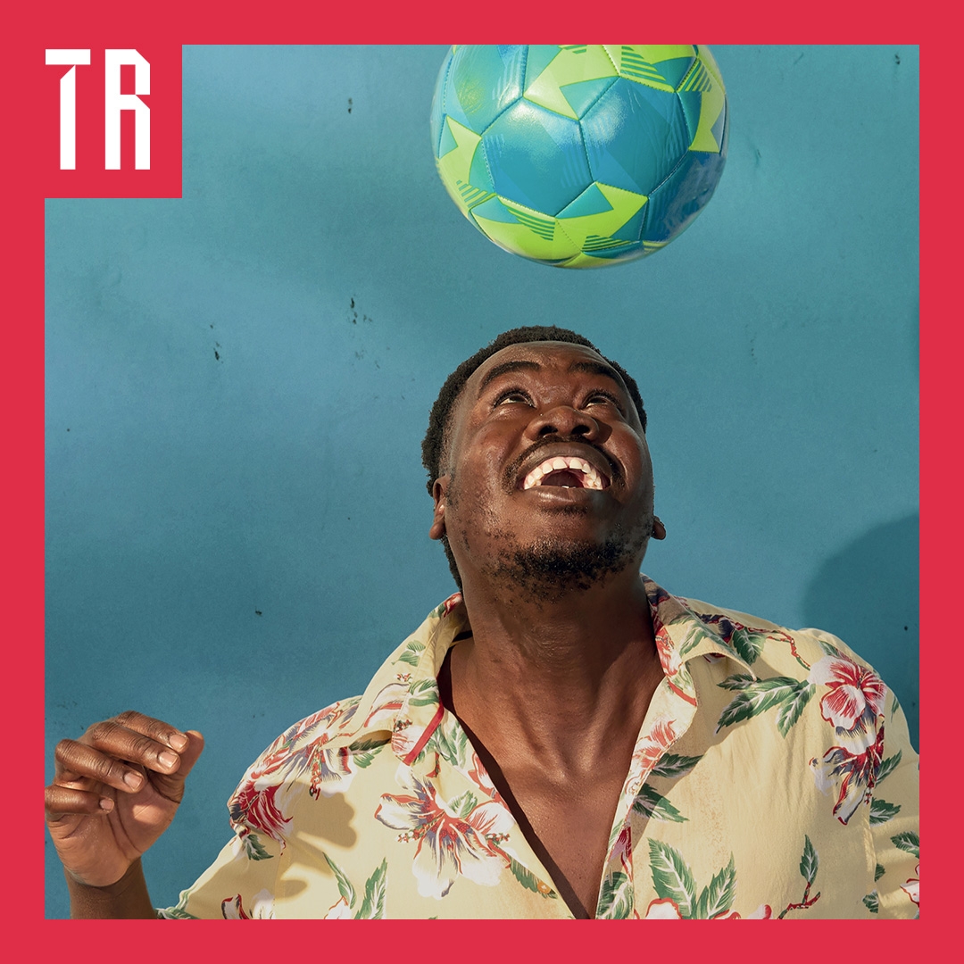 An African man wearing a tropical shirt smile as he looks at a soccer ball suspended mid air